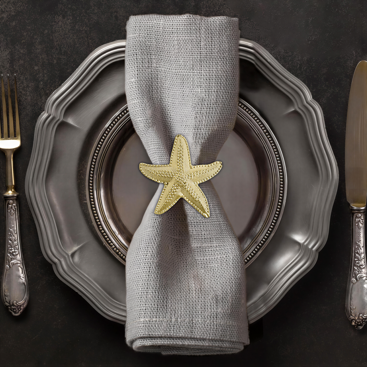 A silver dinner plate with a gold starfish napkin ring, silverware, and a dark background.