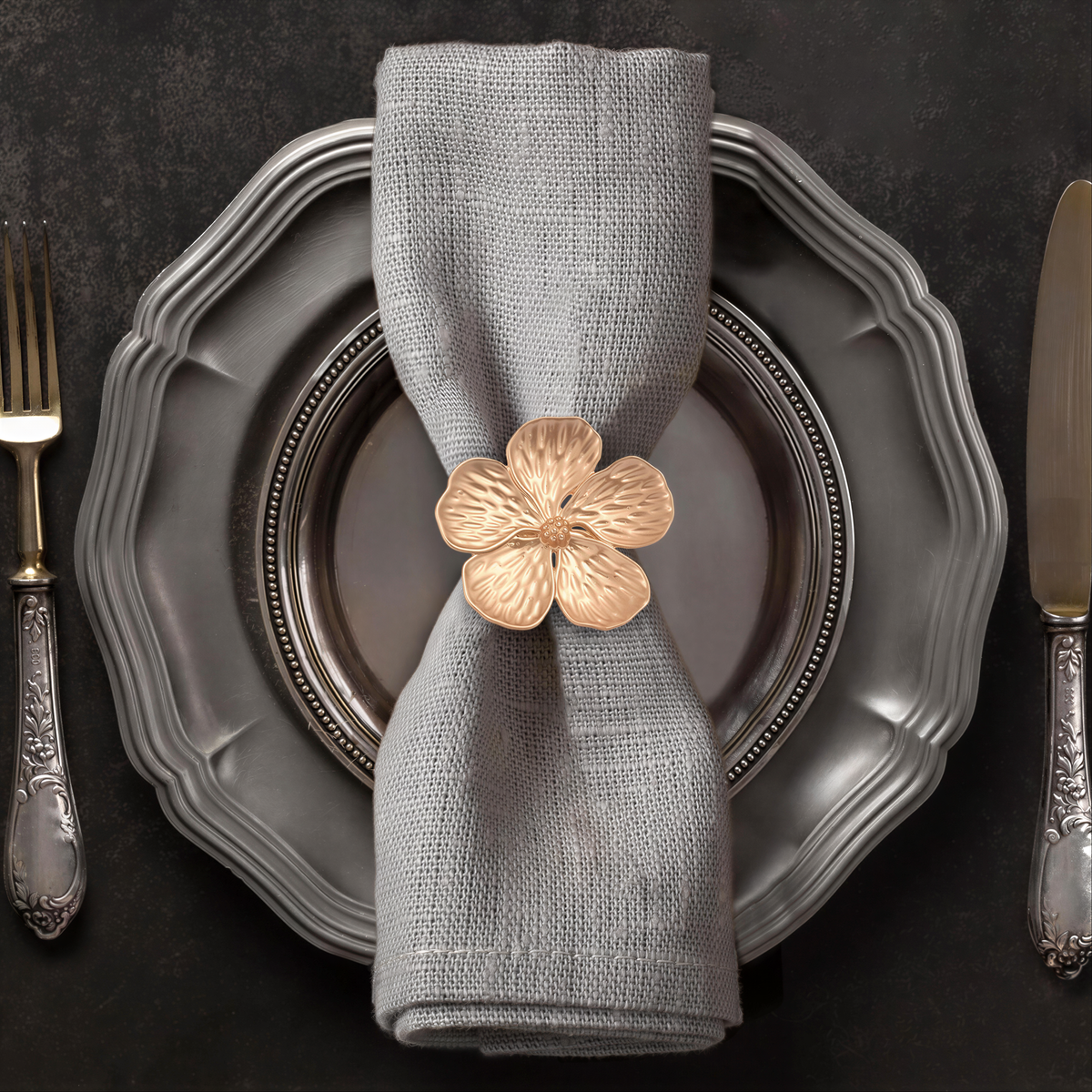 A neatly arranged place setting with a gray napkin, a silver plate, and a gold flower-shaped napkin ring.