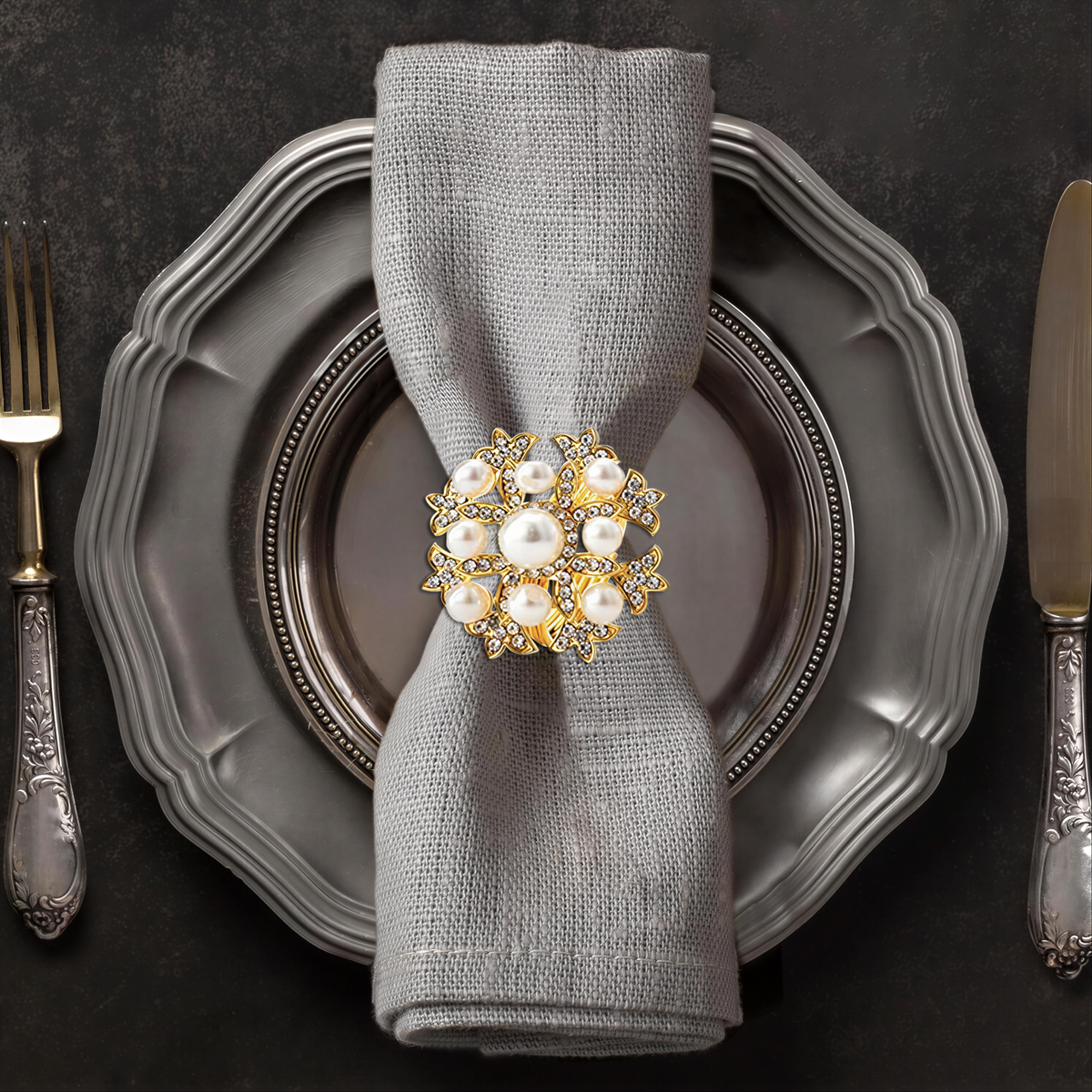 A silver dinner plate with a decorative napkin ring featuring a large, ornate piece of jewelry, likely a brooch or pendant, placed on top of it.