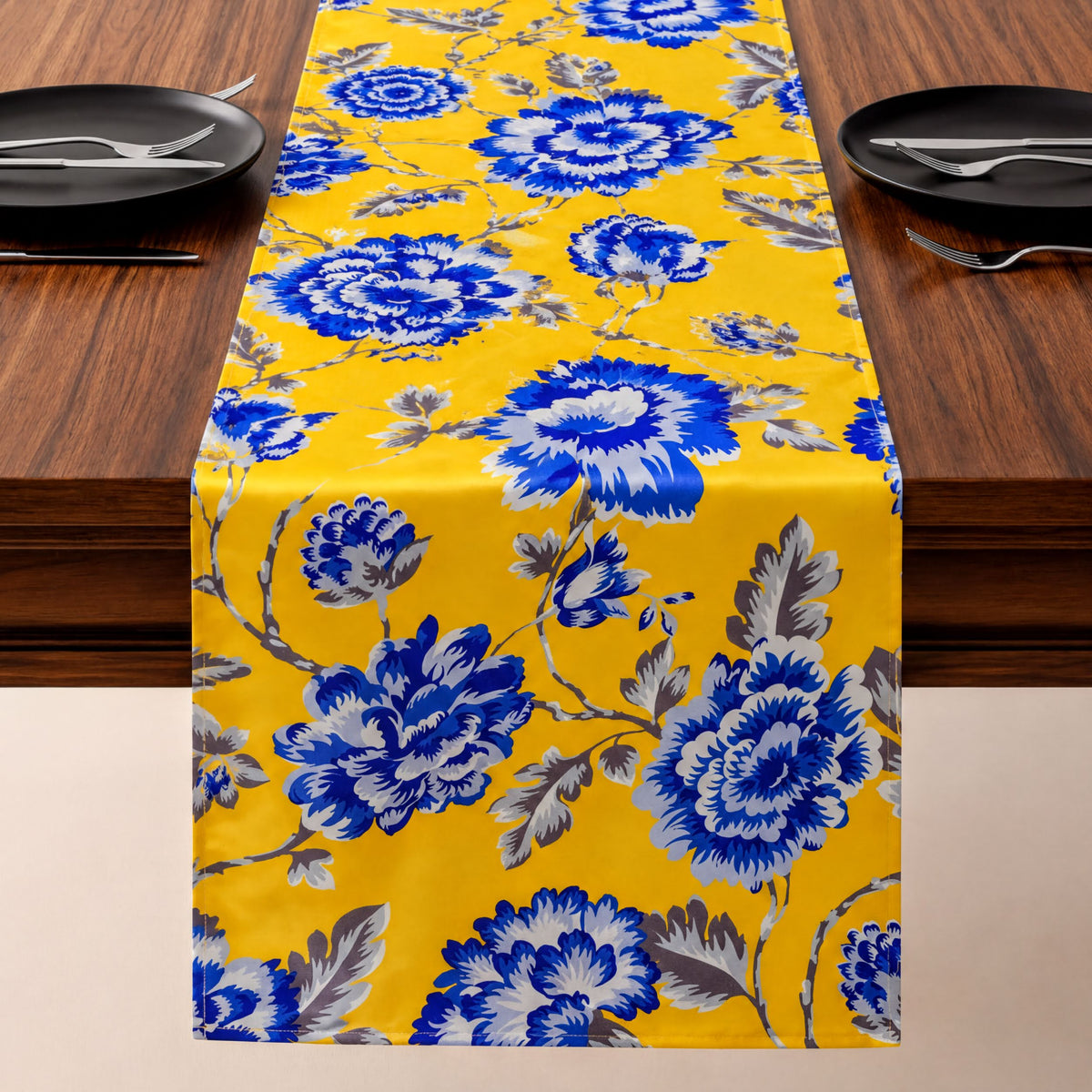 a table runner with a yellow floral print, placed on a wooden table. The runner is adorned with blue and white flowers, creating a vibrant and eye-catching design. The table is set with black plates and silverware, ready for a meal.