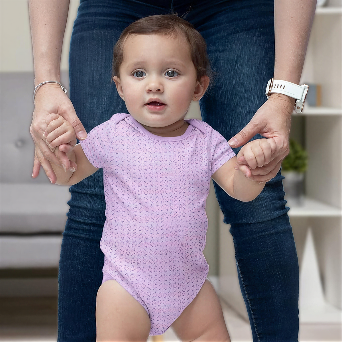 A baby girl wearing a purple onesie is being held by an adult, who is standing behind her.