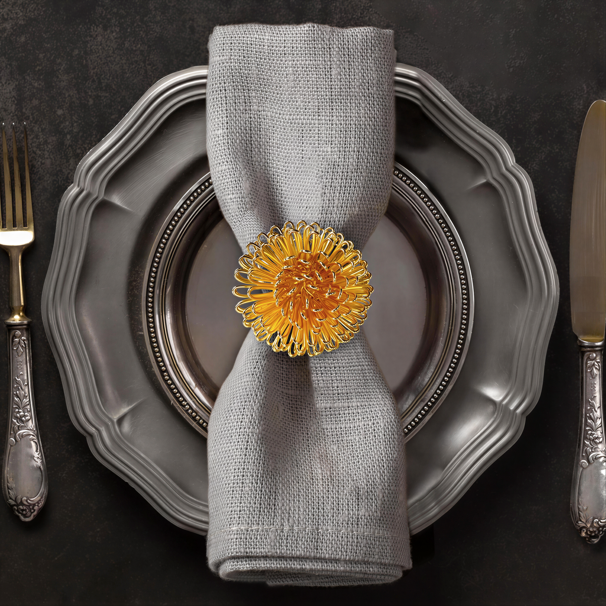 A silver dinner plate with a decorative napkin ring featuring a yellow flower, accompanied by a fork and knife on a dark background.