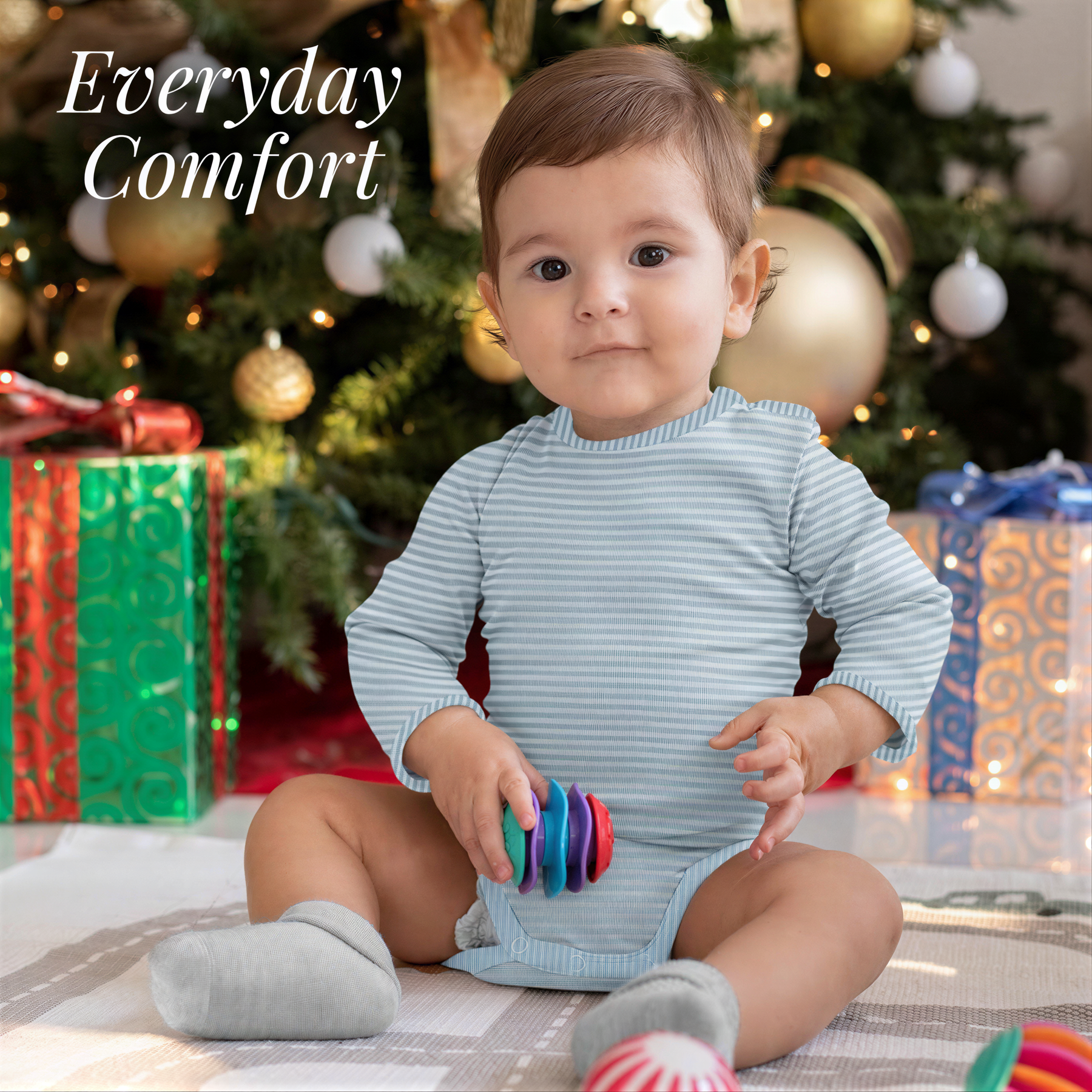 A smiling baby in a striped onesie sits on the floor, holding a colorful toy, with a Christmas tree and wrapped gifts in the background.