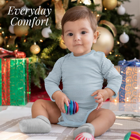 A smiling baby in a striped onesie sits on the floor, holding a colorful toy, with a Christmas tree and wrapped gifts in the background.
