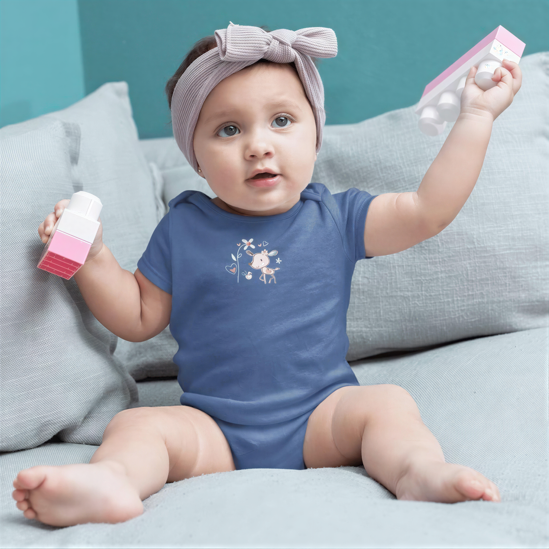 A baby girl wearing a blue onesie and a gray headband is sitting on a gray couch, holding a pink and white toy.