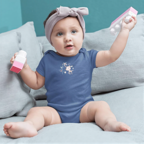 A baby girl wearing a blue onesie and a gray headband is sitting on a gray couch, holding a pink and white toy.