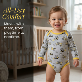 A smiling baby boy wearing a gray and yellow onesie with a truck pattern, standing in a nursery setting.