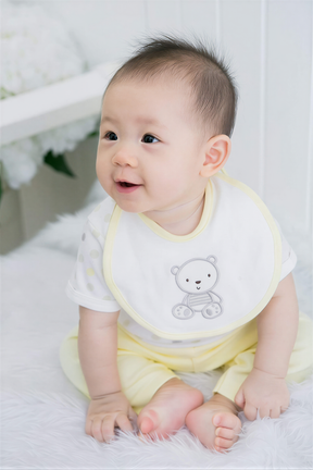 A smiling baby boy wearing a yellow bib with a teddy bear design sits on a white surface.