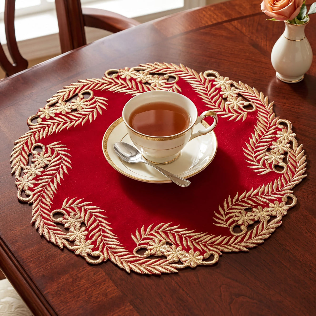 A cup of tea sits on a red and gold place mat, accompanied by a small vase of flowers on a wooden table.
