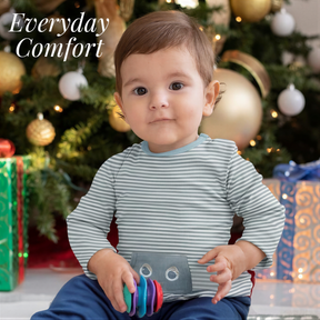 A young child sitting in front of a Christmas tree, holding a toy, with a gift box and ornaments visible in the background.