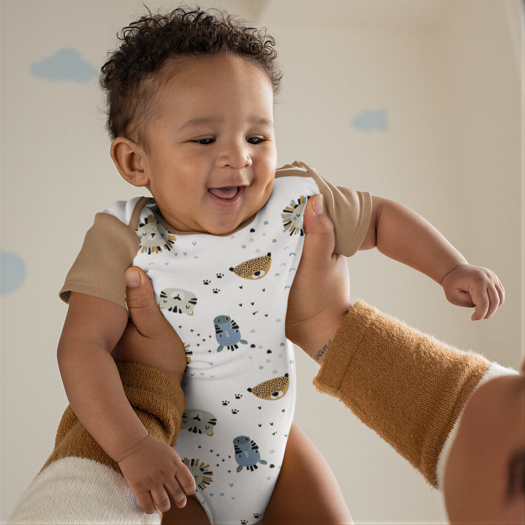 A smiling baby wearing a white onesie with animal prints is being held by an adult's arms.