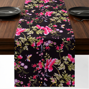 a table runner with a floral pattern in shades of pink and green, placed on a wooden table. The runner is adorned with small pink flowers and green leaves, creating a visually appealing and decorative touch to the table setting.