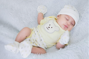 A sleeping baby wearing a yellow outfit with a white bear design, lying on a white fur-covered surface.