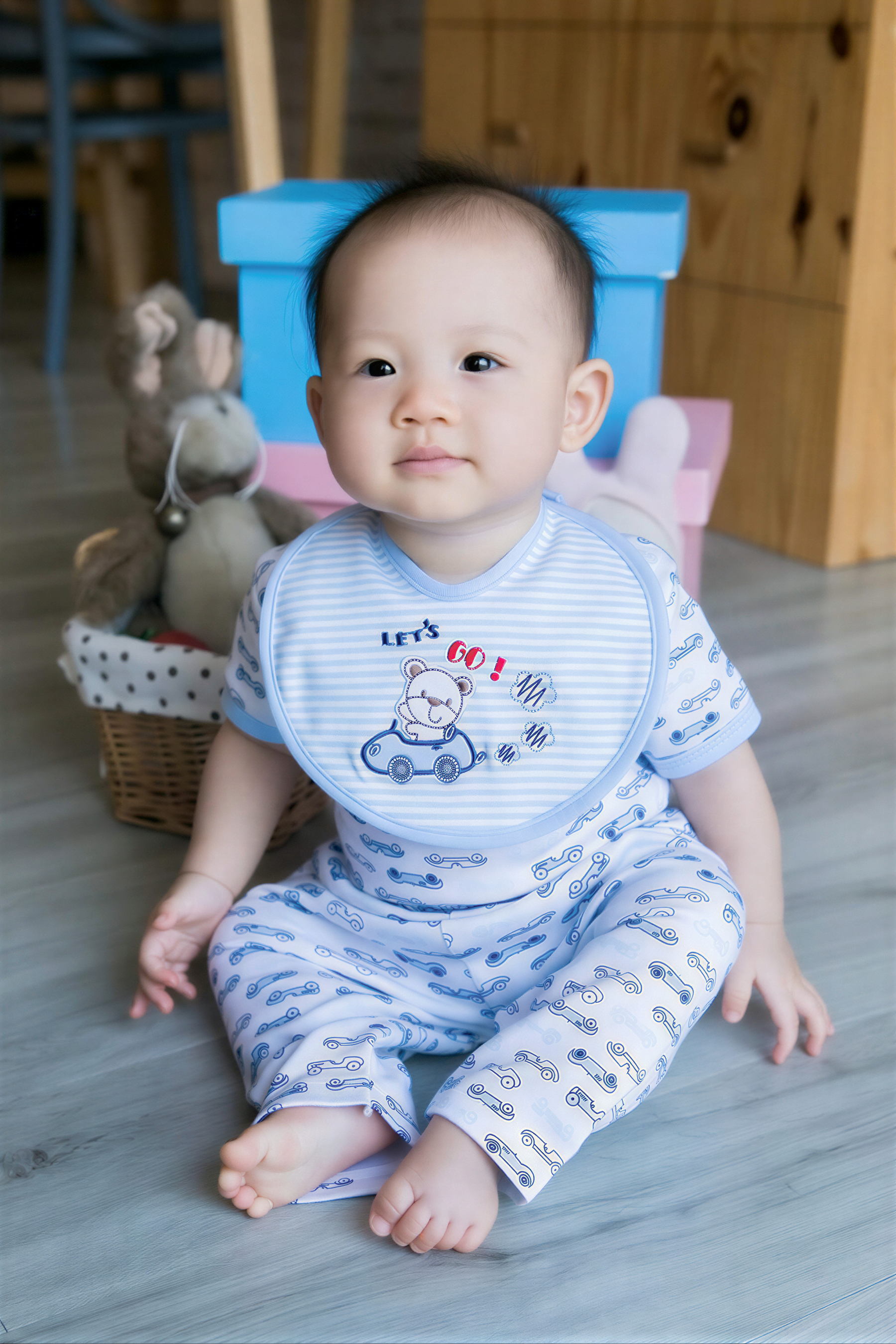 A smiling baby boy wearing a bib with a cartoon character sits on the floor, surrounded by toys and a blue box.