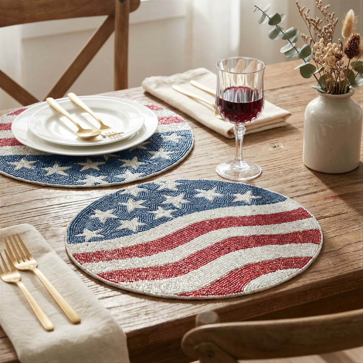 a table setting with a white plate, a beaded placemat with an American flag design, a wine glass, a vase with flowers, and a set of gold cutlery.