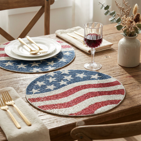 a table setting with a white plate, a beaded placemat with an American flag design, a wine glass, a vase with flowers, and a set of gold cutlery.