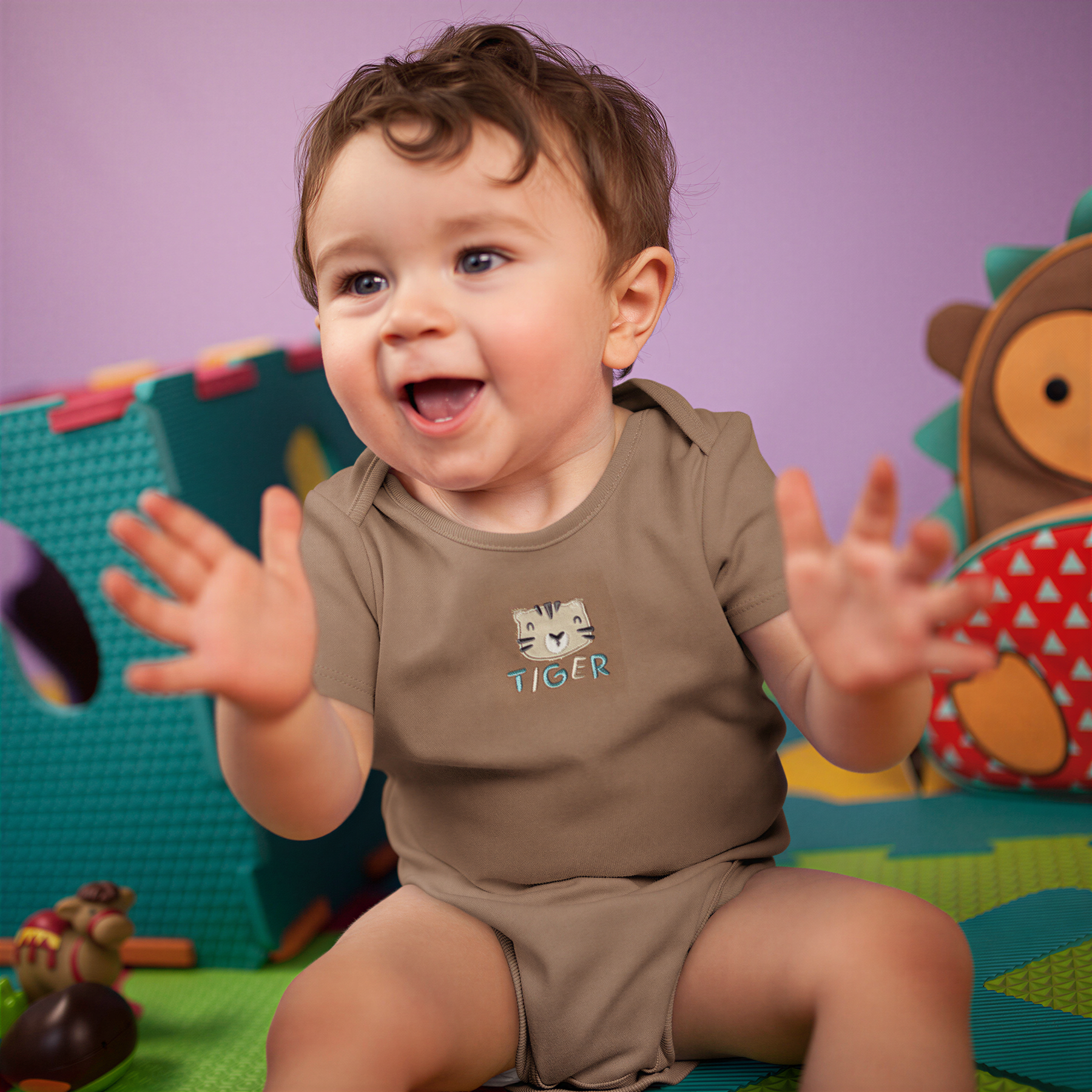 A happy baby boy wearing a brown shirt with a tiger design, sitting on a colorful play mat and smiling.