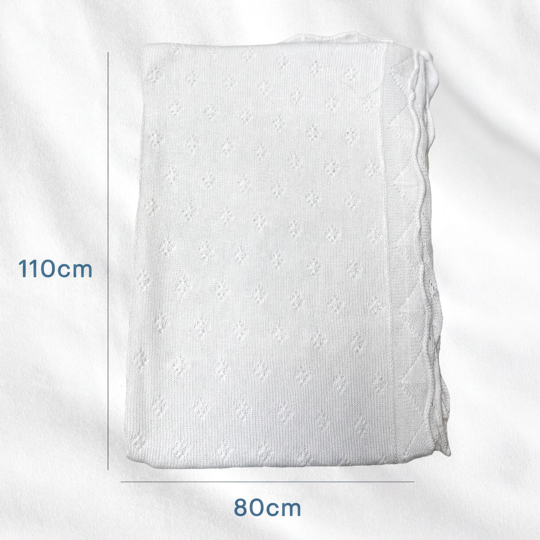 a white knitted blanket or quilt with a pattern of small holes or dots. The blanket is folded and placed on a white surface, with measurements provided in centimeters.