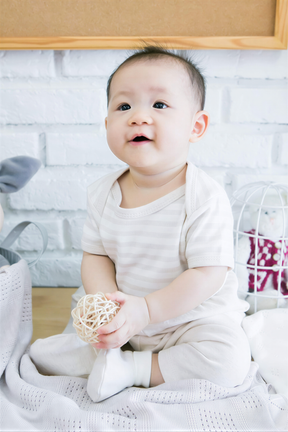 A smiling baby in a white outfit sits on a blanket, holding a ball of string.