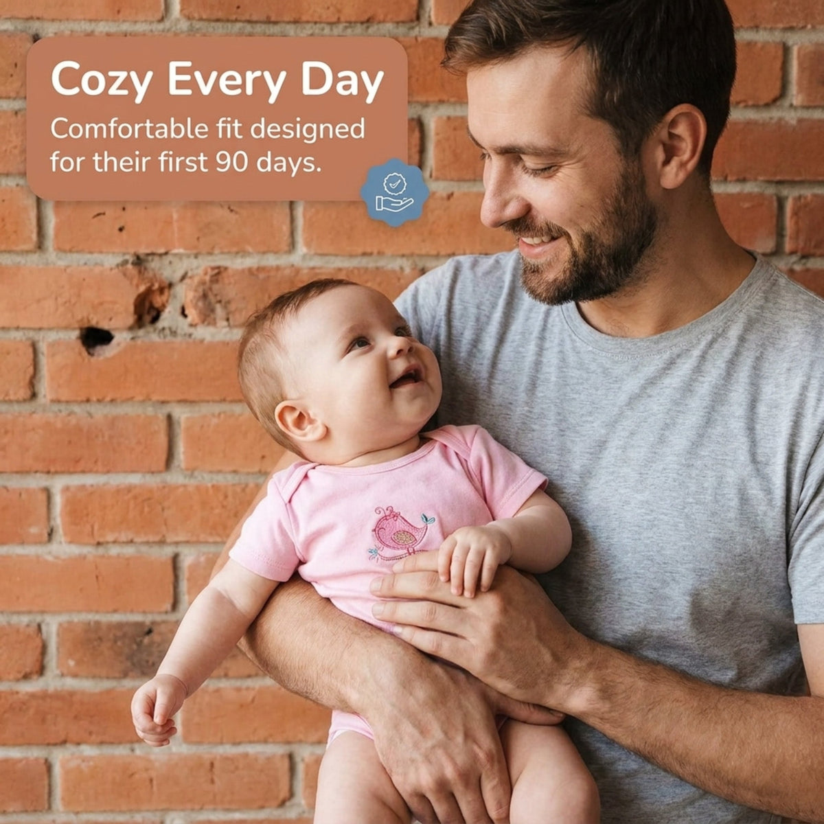 A man in a gray shirt is holding a smiling baby in a pink onesie, with a brick wall in the background.