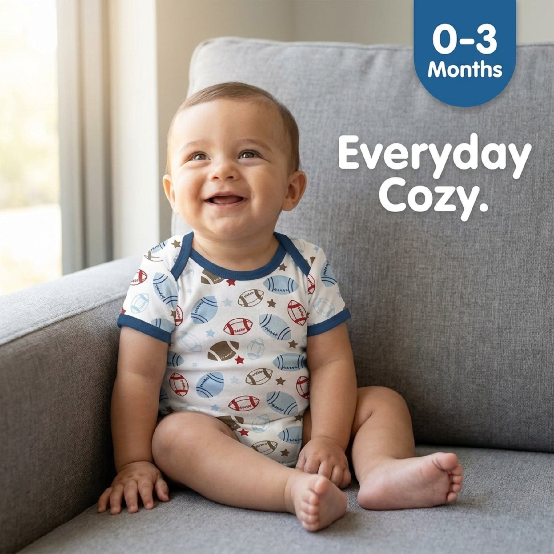 A smiling baby wearing a white onesie with a football pattern sits on a gray couch.