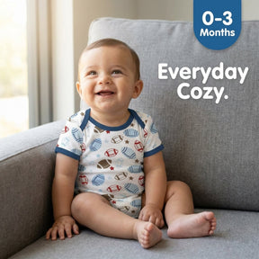 A smiling baby wearing a white onesie with a football pattern sits on a gray couch.