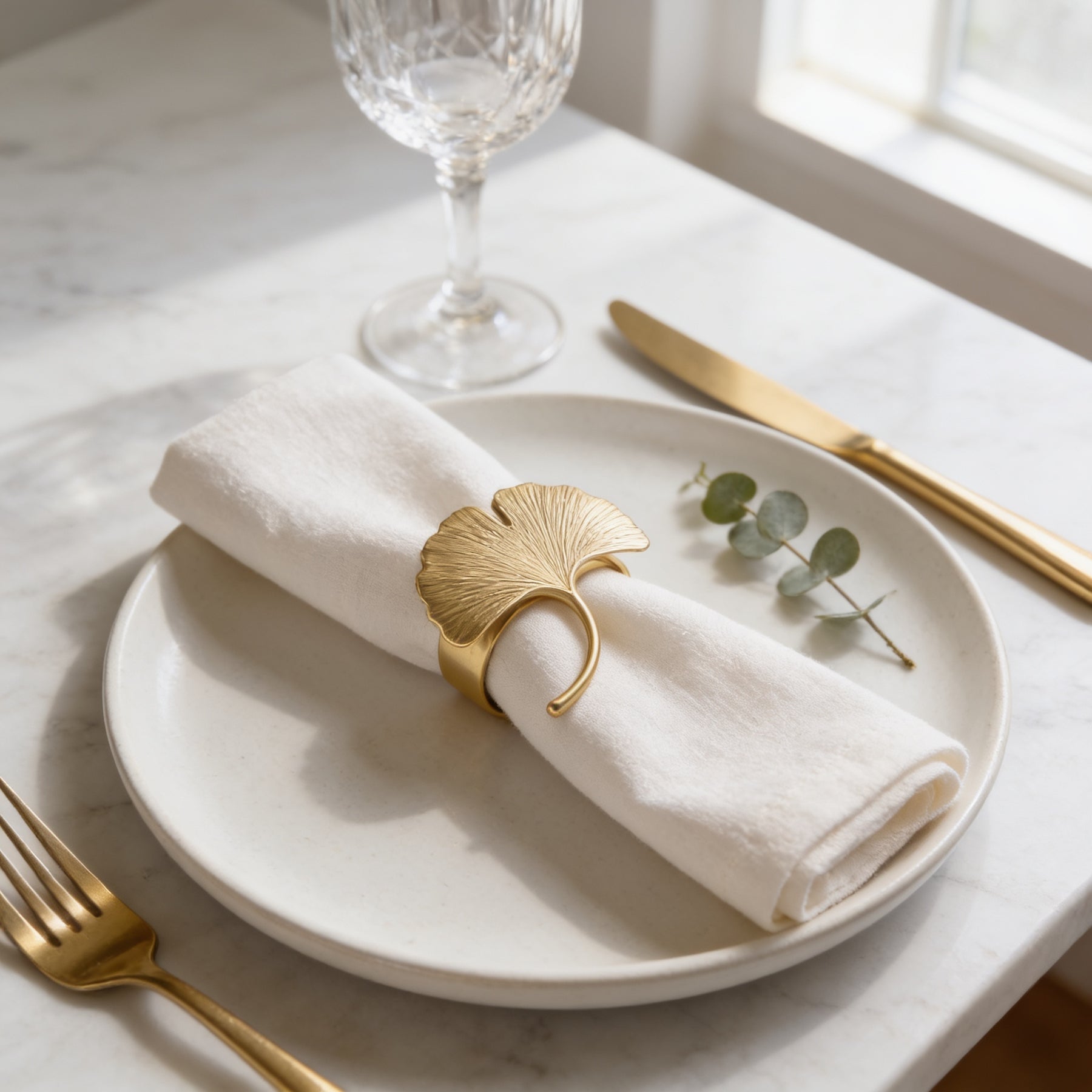 A white plate with a gold napkin ring and a small sprig of greenery, accompanied by a fork and knife, on a marble table.