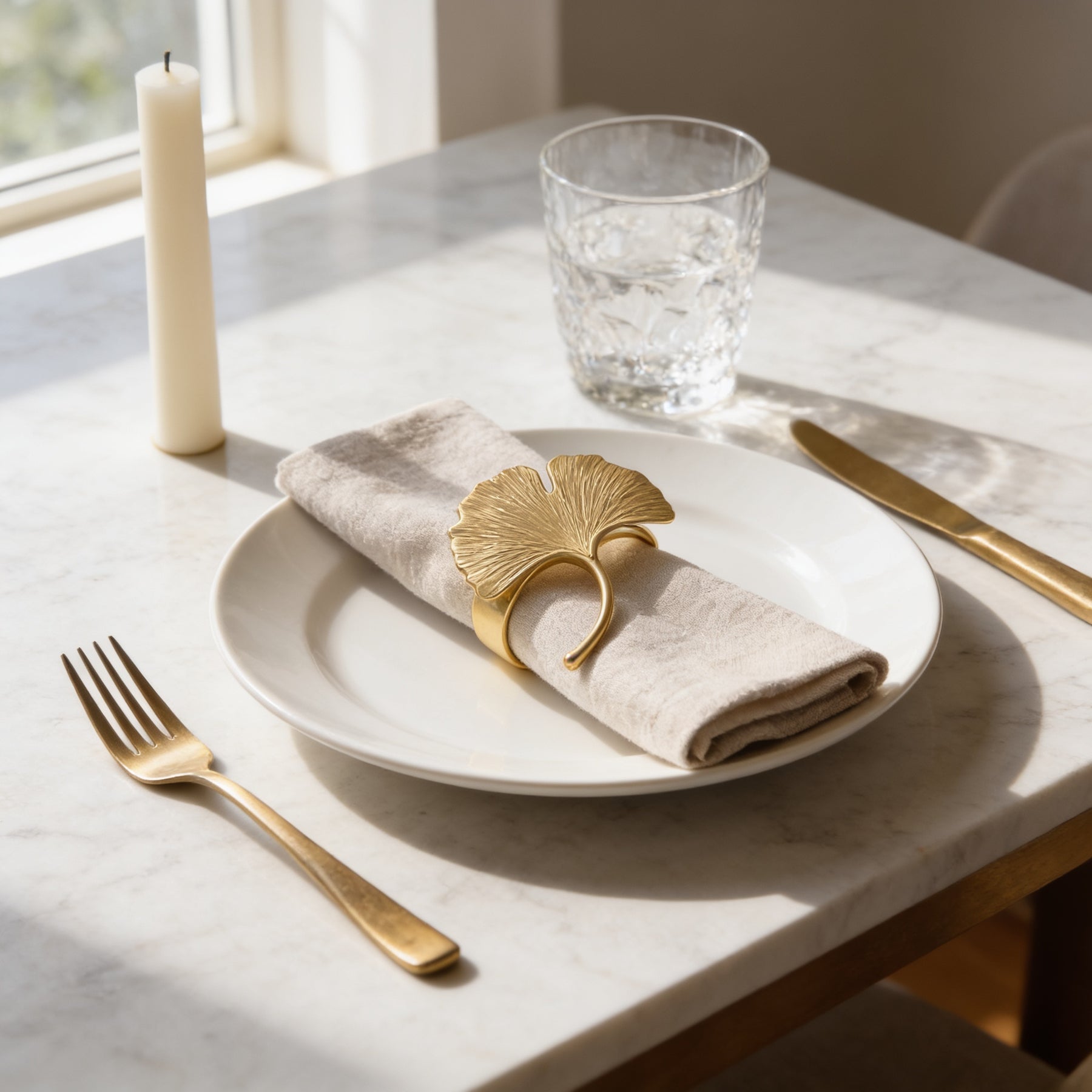 A white plate with a gold napkin ring and a candle on a marble table, accompanied by a fork and knife.