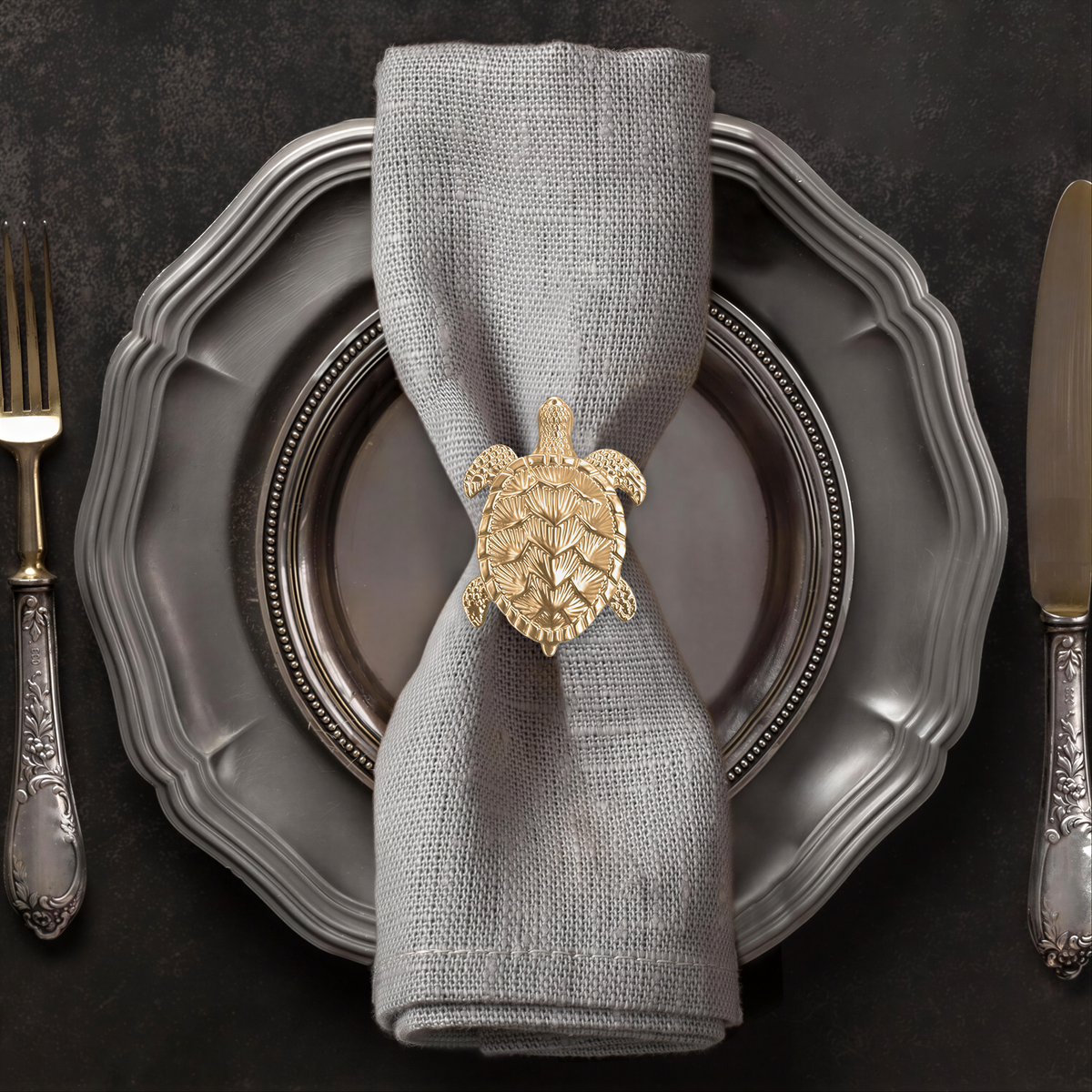A neatly arranged place setting with a silver plate, a folded napkin, and a decorative gold turtle-shaped ring on the napkin ring.