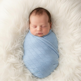 A newborn baby is peacefully sleeping, swaddled in a light blue blanket and resting on a soft, fluffy white surface.