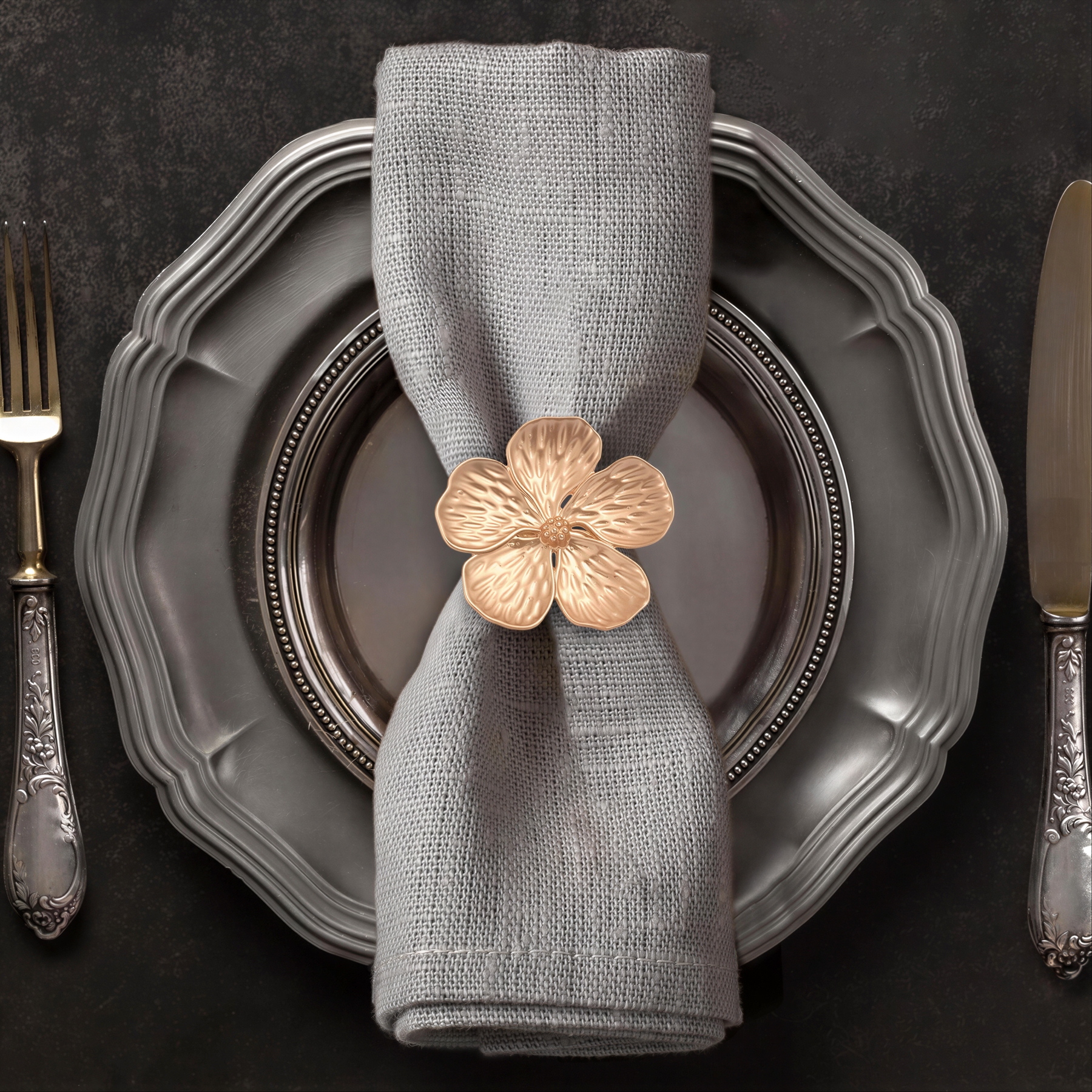 A neatly arranged place setting with a gray napkin, a silver plate, and a gold flower-shaped napkin ring.