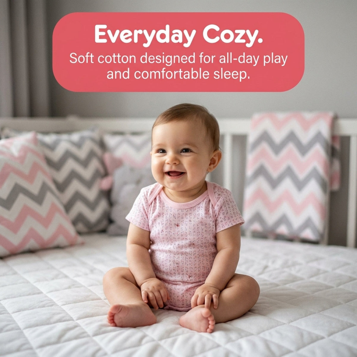 A smiling baby girl sits on a bed, wearing a pink onesie.