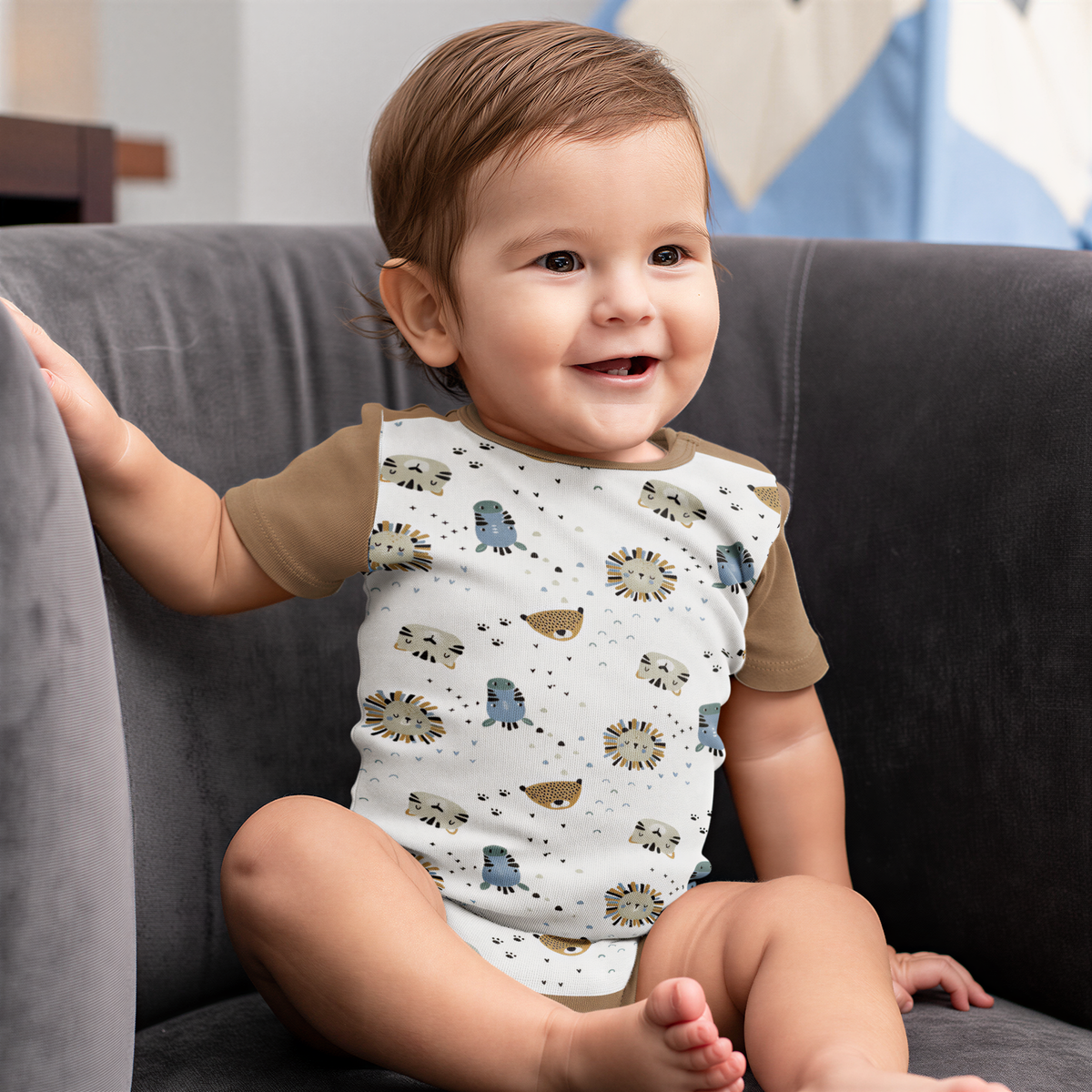 A smiling baby wearing a white onesie with animal prints sits on a gray couch.