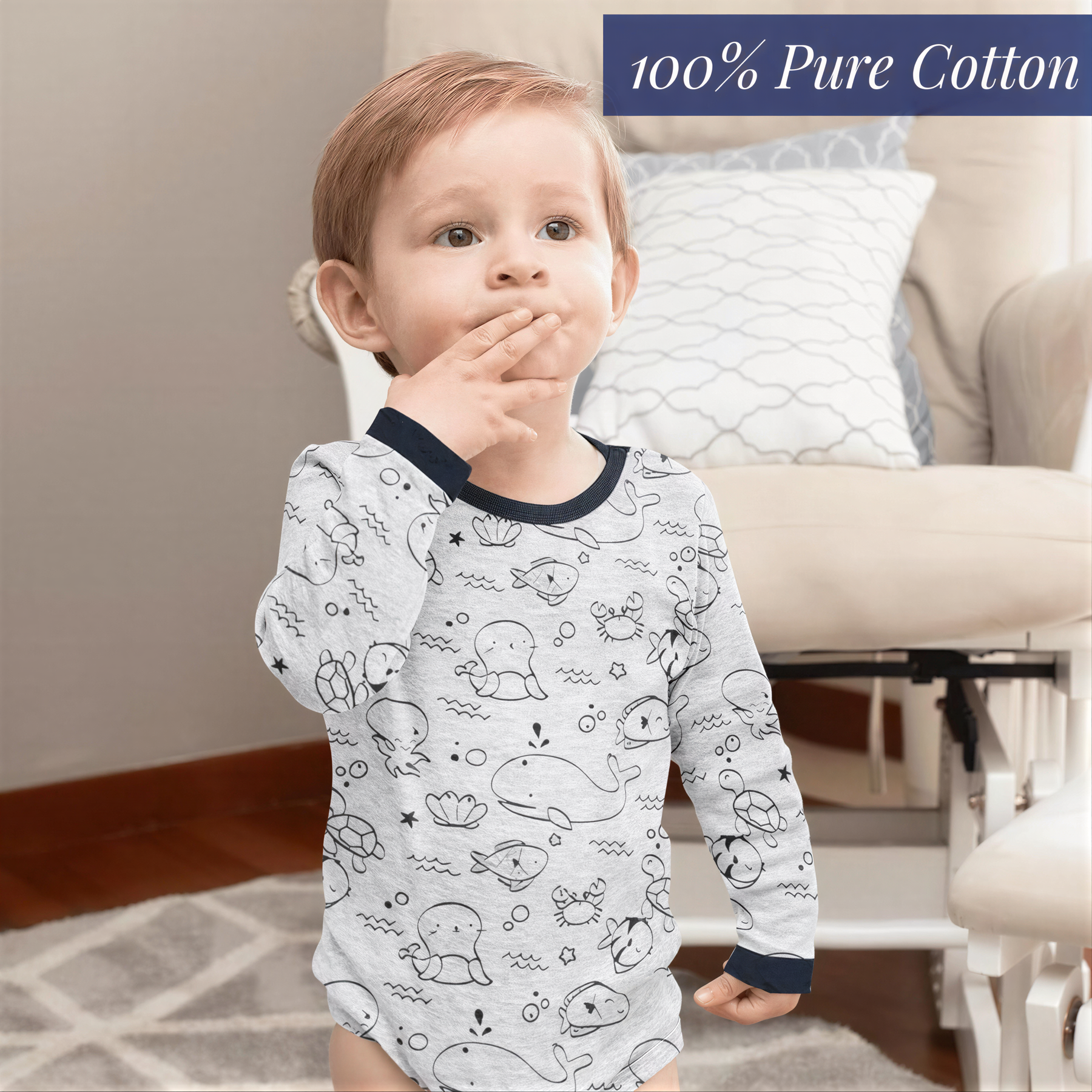 A young child wearing a long-sleeved shirt with a cartoon fish design, standing in a room with a beige chair and a pillow.