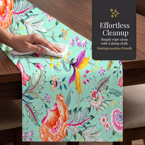 a hand wiping a table runner with a damp cloth, which has a colorful floral pattern. The table is set with a green table runner, and there is a sign in the background that reads "Effortless Cleanup" and "Simply wipe clean with a damp cloth. Washing machine friendly".