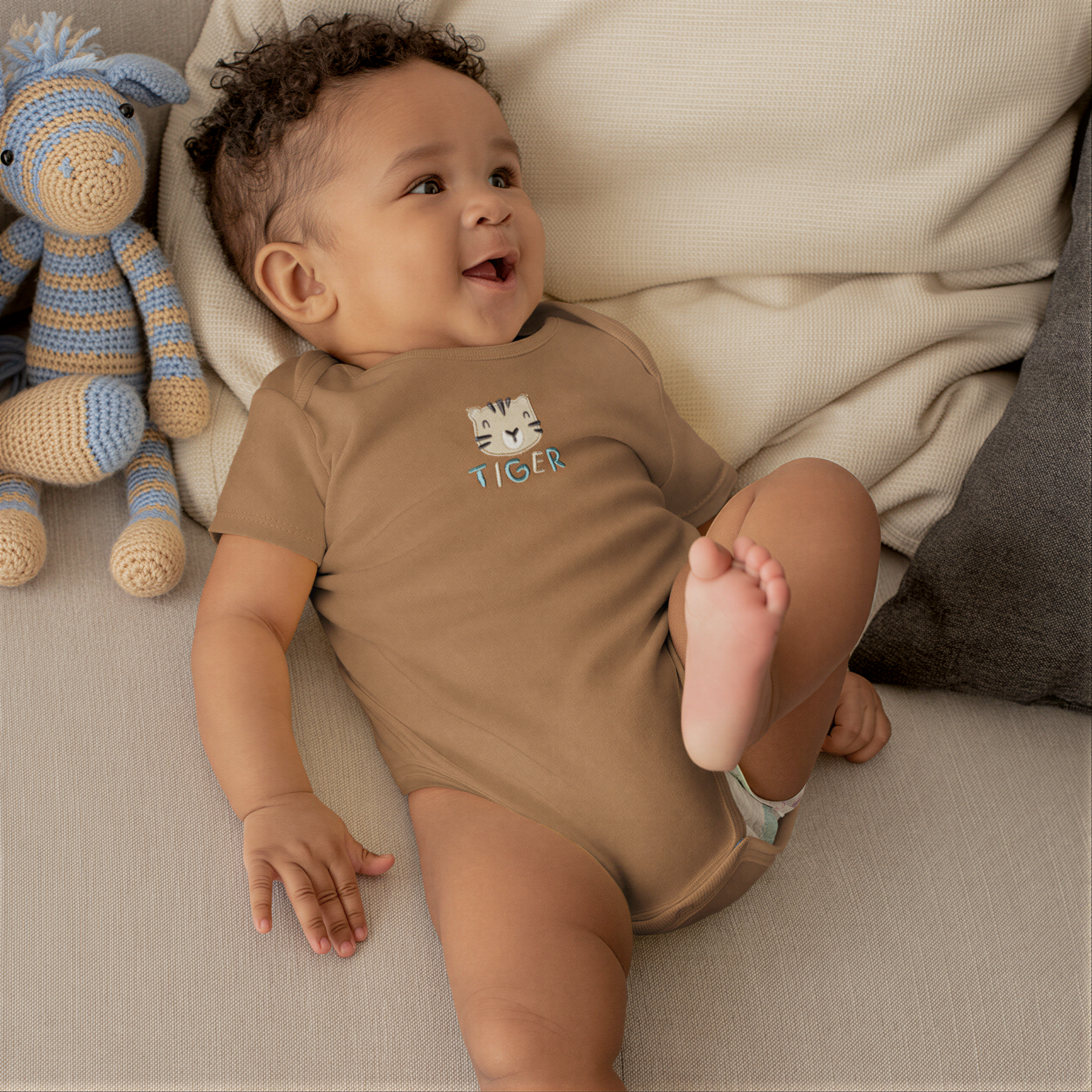 A smiling baby boy wearing a brown onesie with a tiger design lies on a couch next to a stuffed animal.