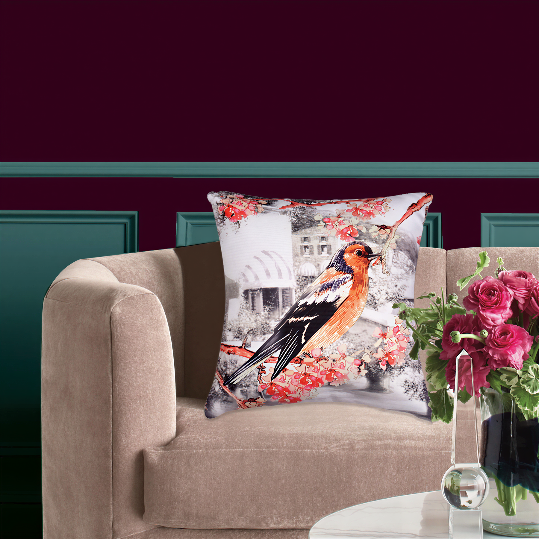 a beige sofa with a decorative pillow featuring a colorful bird design, placed on top of it. Next to the sofa, there is a vase filled with pink flowers, creating a cozy and inviting atmosphere in the living room.
