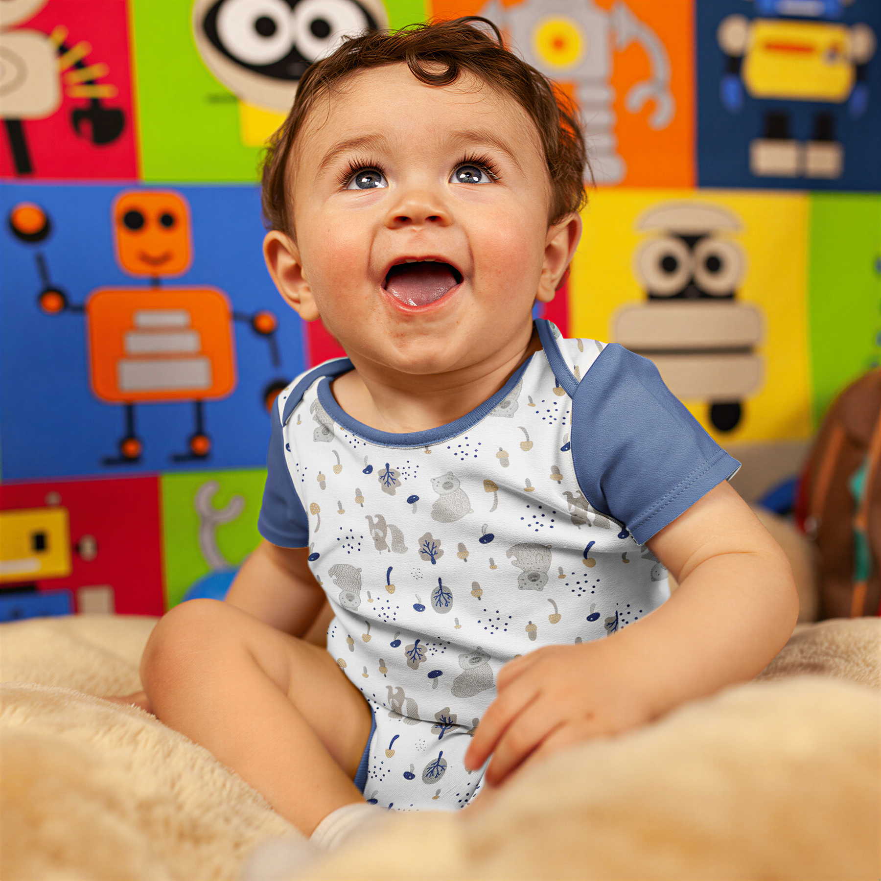 A smiling baby boy sitting on a bed, wearing a white and blue patterned onesie.