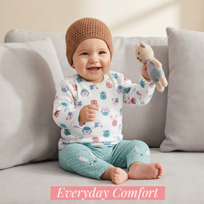 A smiling baby in a white onesie with animal prints and a brown knit cap sits on a gray couch, holding a small stuffed animal.