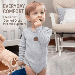 A young child wearing a gray onesie with a hedgehog design, standing in a nursery setting with a rocking chair and a stuffed animal.