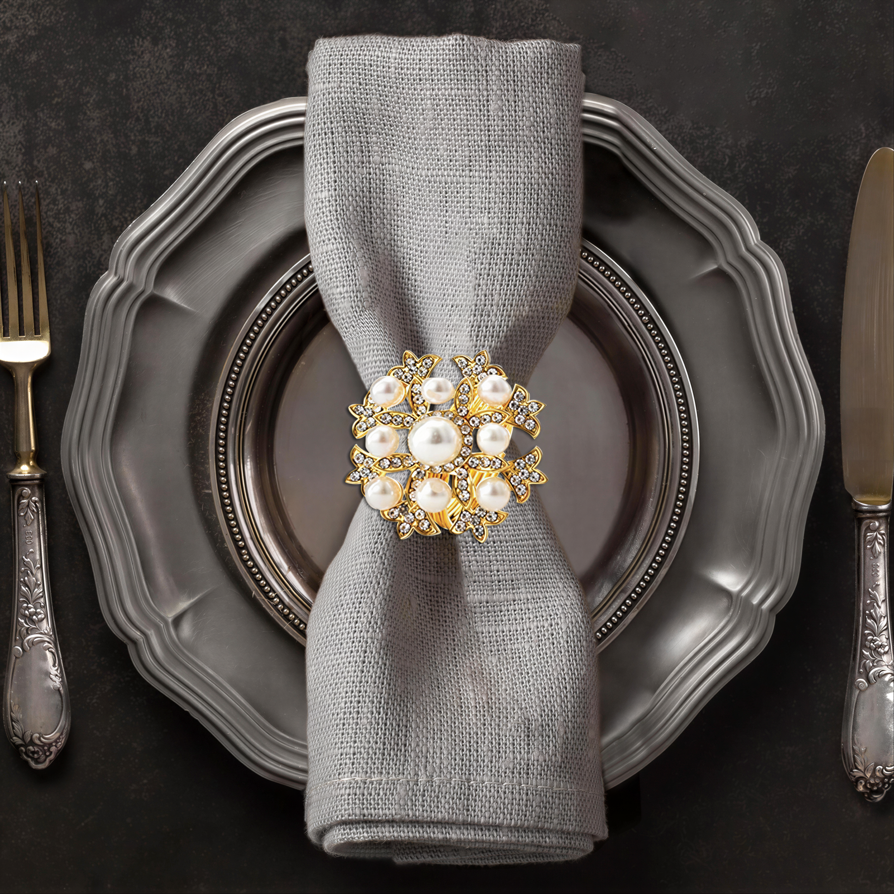A silver dinner plate with a decorative napkin ring featuring a large, ornate piece of jewelry, likely a brooch or pendant, placed on top of it.