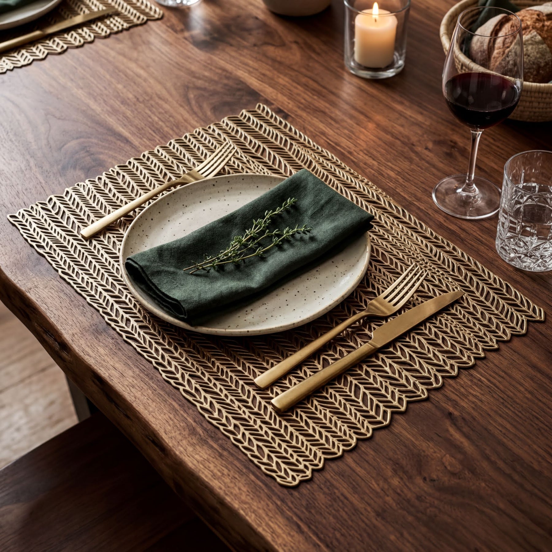 a table setting with a white plate, a green napkin, and a gold fork and knife. The table is set with a woven placemat, and there are glasses of wine and a candle on the table.