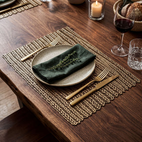 a table setting with a white plate, a green napkin, and a gold fork and knife. The table is set with a woven placemat, and there are glasses of wine and a candle on the table.