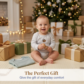 A smiling baby sitting on the floor surrounded by wrapped gifts, with a Christmas tree in the background.