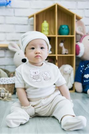 A baby boy wearing a white outfit and bib sits on the floor in front of a wooden toy house, surrounded by various stuffed animals.