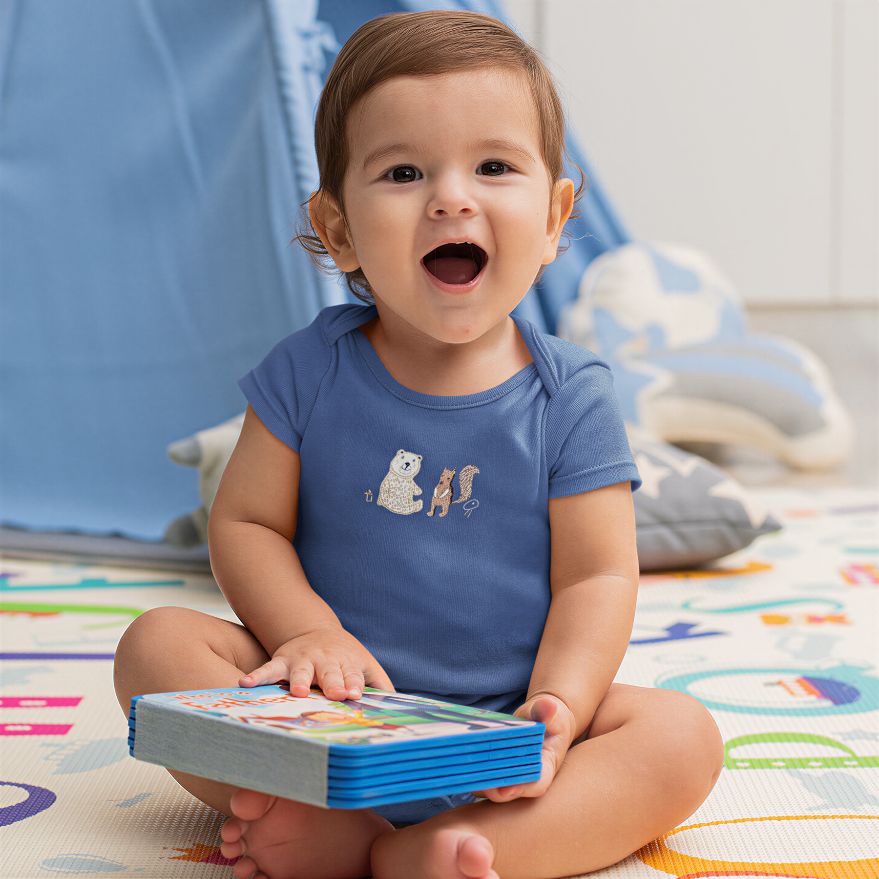 A smiling baby boy sitting on the floor, holding a book and looking at the camera.