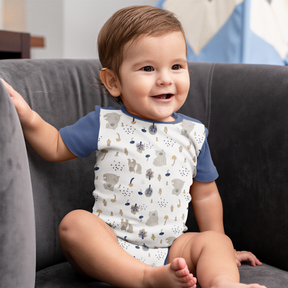 A smiling baby boy wearing a white and blue patterned onesie sits on a gray couch.
