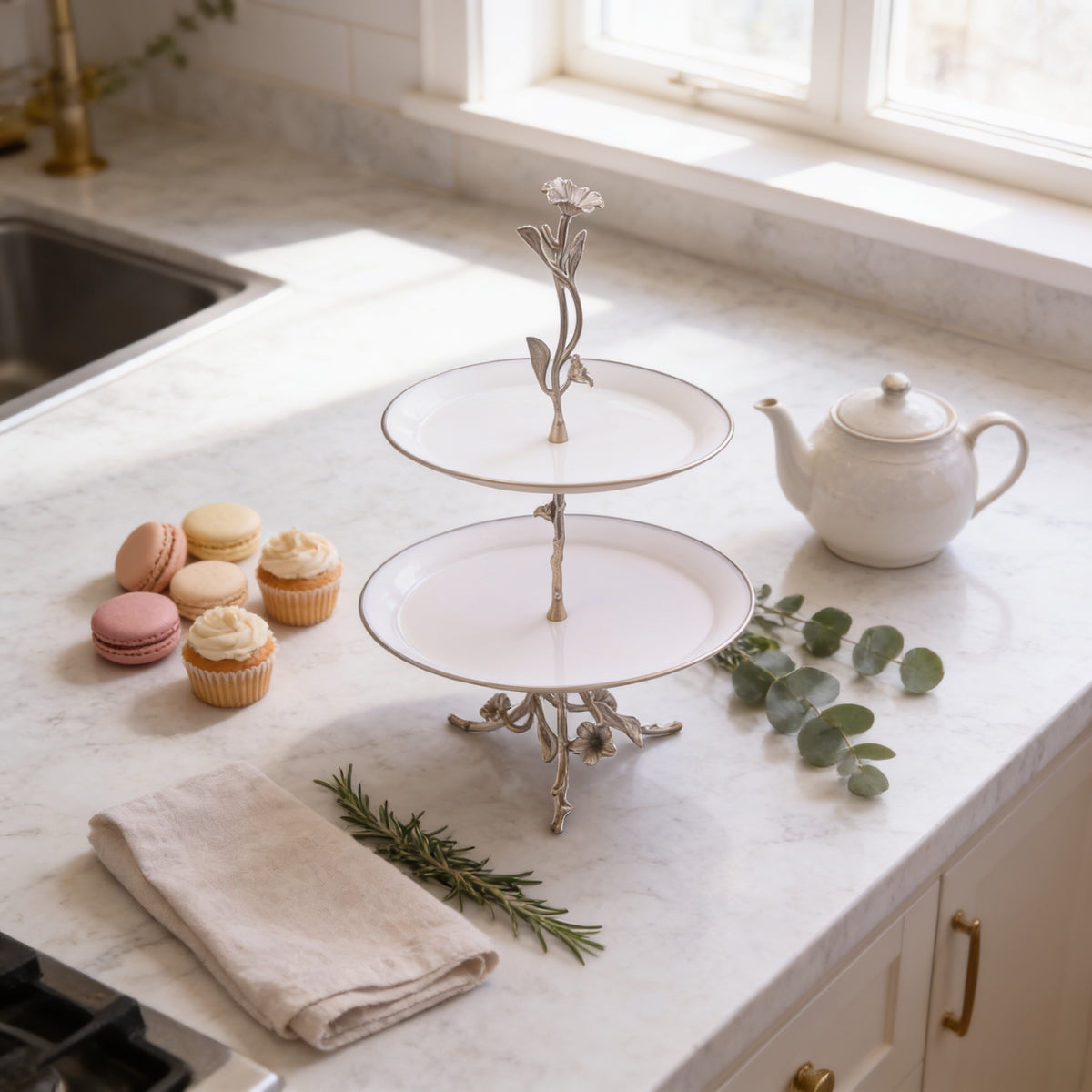 a kitchen counter with a two-tiered cake stand holding a teapot and a set of cupcakes. The counter also has a tea pot, a teacup, and some greenery, creating a cozy and inviting atmosphere.
