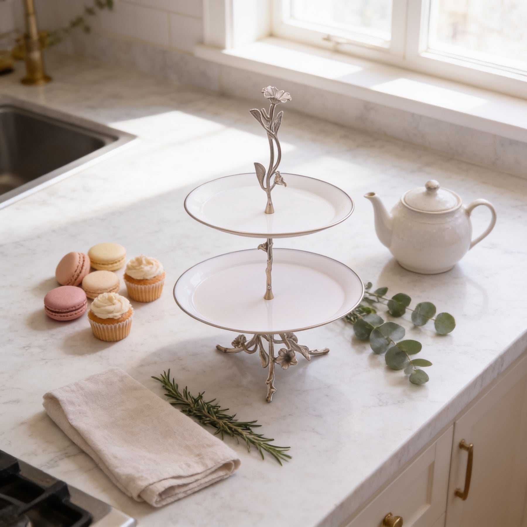 a kitchen counter with a two-tiered cake stand holding a teapot and a set of cupcakes. The counter also has a tea pot, a teacup, and some greenery, creating a cozy and inviting atmosphere.