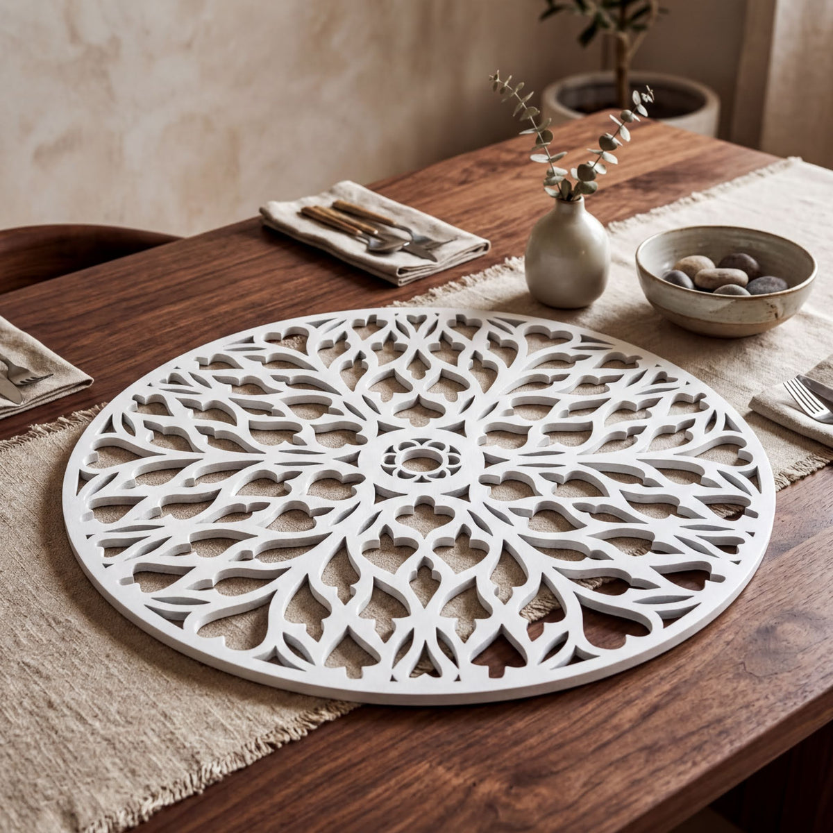 a wooden table with a decorative white placemat, a bowl of rocks, a vase with greenery, and a set of silverware.